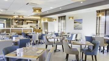 A modern restaurant with neatly arranged tables and chairs, a bar area with shelves of bottles, and pendant lighting.