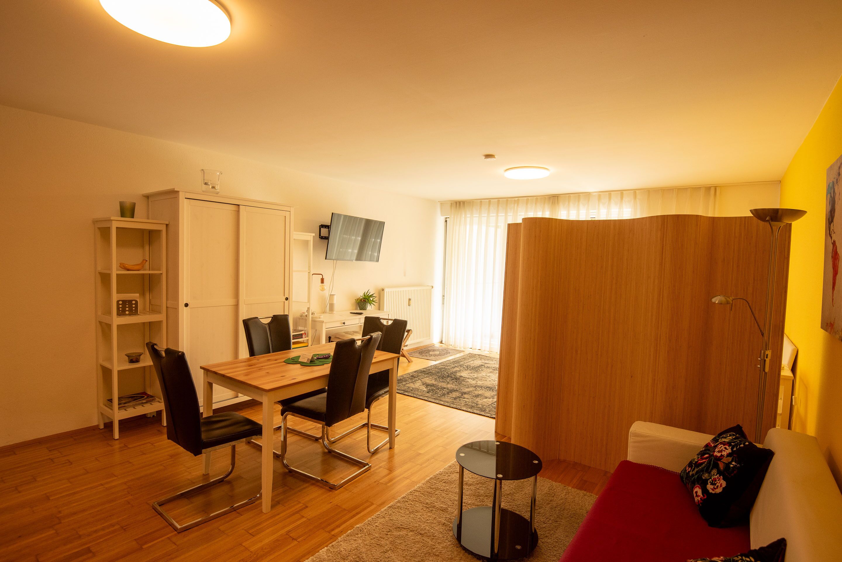 Un salon moderne avec un coin repas, comprenant une table à manger en bois, des chaises noires, un canapé rouge et un téléviseur fixé au mur.