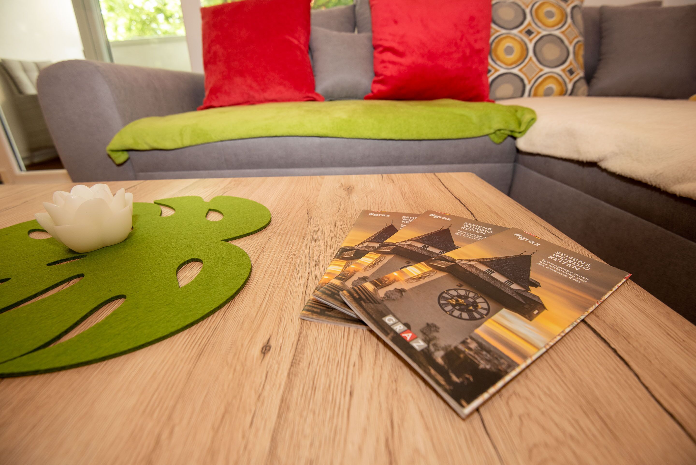 Una mesa de madera con un adorno verde y revistas, un sofá gris con cojines rojos y verdes, y una ventana al fondo.