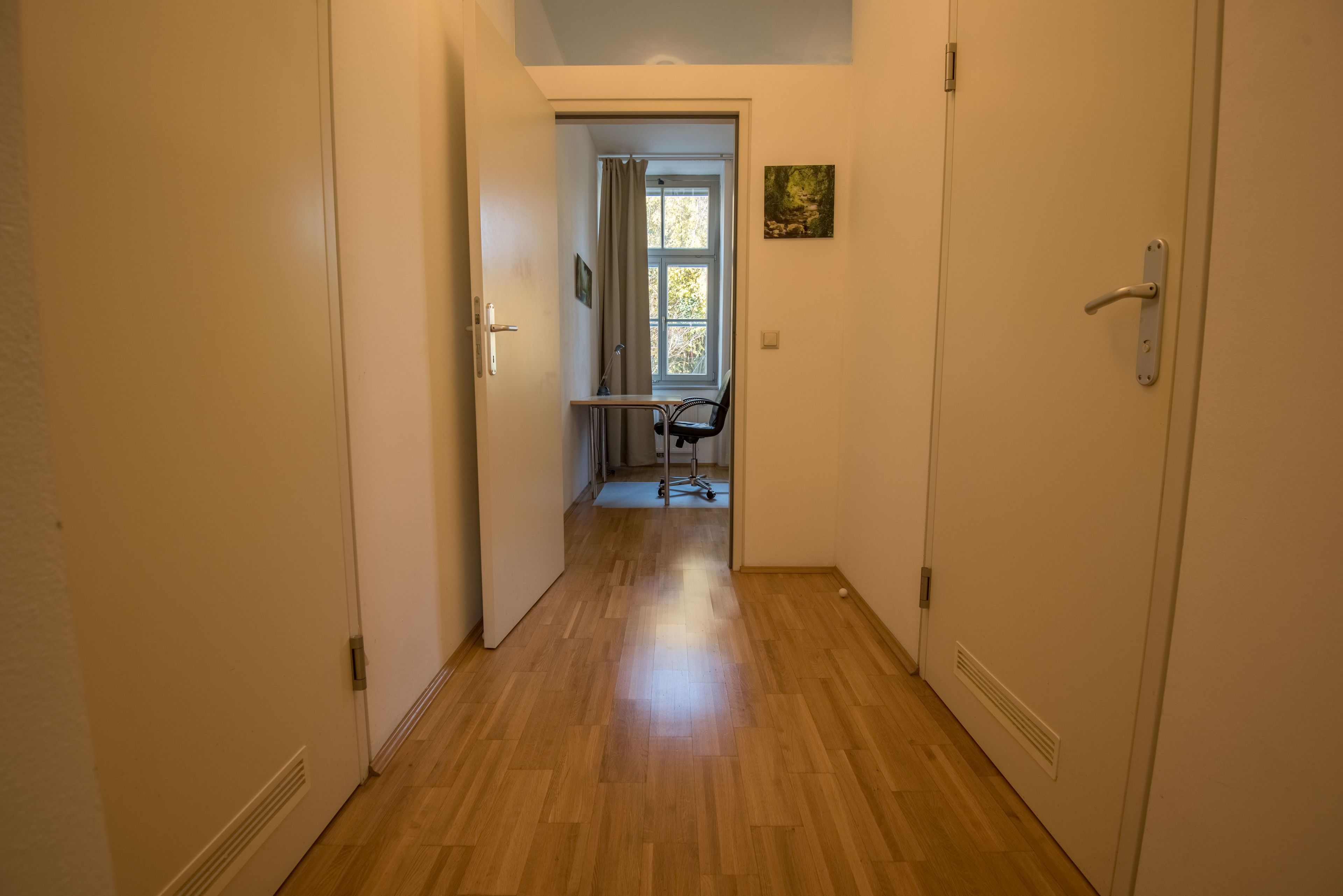 A narrow hallway with wooden flooring leading to a room with a window and a chair.