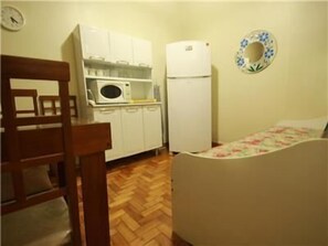 A compact kitchen with white cabinets, a microwave, and a refrigerator. There is a wooden chair and a bed with a floral bedspread. - GoHouse Copacabana 1203 (Rio de Janeiro)