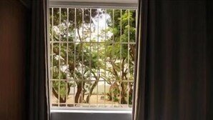 A room with a window featuring metal bars, curtains, and a view of trees and a building outside. - GoHouse Copacabana 204 B (Rio de Janeiro)