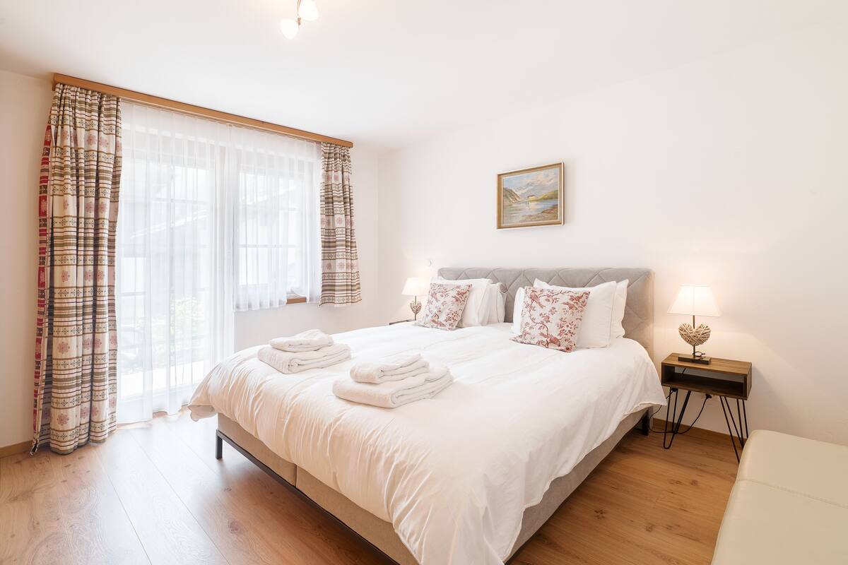 A neatly made bed with white linens and folded towels, a bedside table with a lamp, and a painting on the wall.