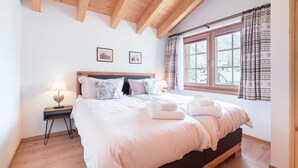 Quarto com teto de madeira, cama grande com roupa de cama branca, mesa de cabeceira com abajur e janela com cortinas estampadas.