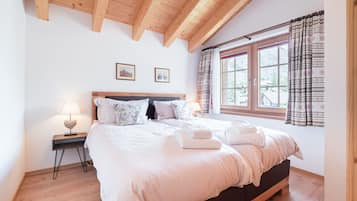 A bedroom with a wooden ceiling, a large bed with white bedding, a nightstand with a lamp, and a window with patterned curtains.