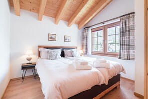 Ein Schlafzimmer mit Holzboden, einem großen Bett mit weißer Bettwäsche, einem Nachttisch mit Lampe und einem Fenster mit gemusterten Vorhängen.