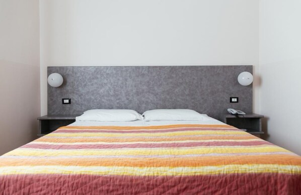 Una cama bien hecha con un edredón a rayas, dos mesitas de noche con lámparas y una pared lisa.