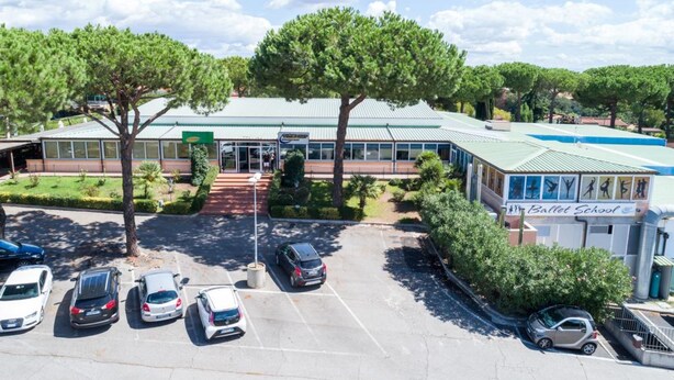 A building with a green roof, a sign for a ballet school, and a parking lot with several cars. - Autohotel (Formello)