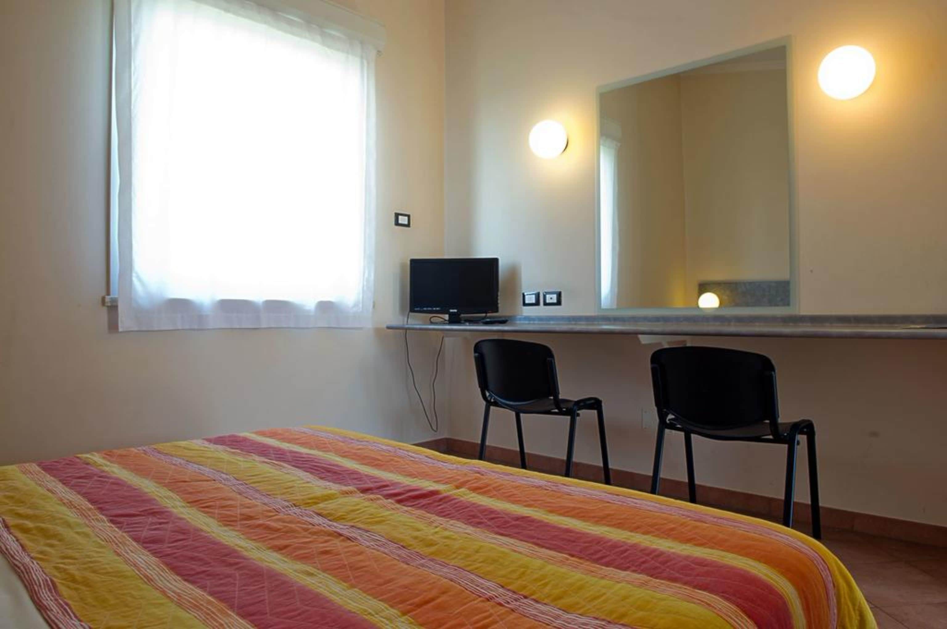 Ein Schlafzimmer mit einem Bett, zwei Stühlen, einem Schreibtisch mit Computer und einem Fenster mit Vorhängen sowie einem Spiegel.
