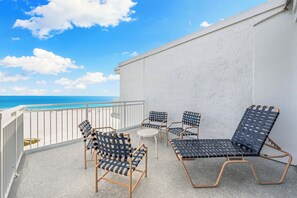 A balcony with a view of the ocean, featuring a white wall, a metal railing, and several wicker chairs and a small table.