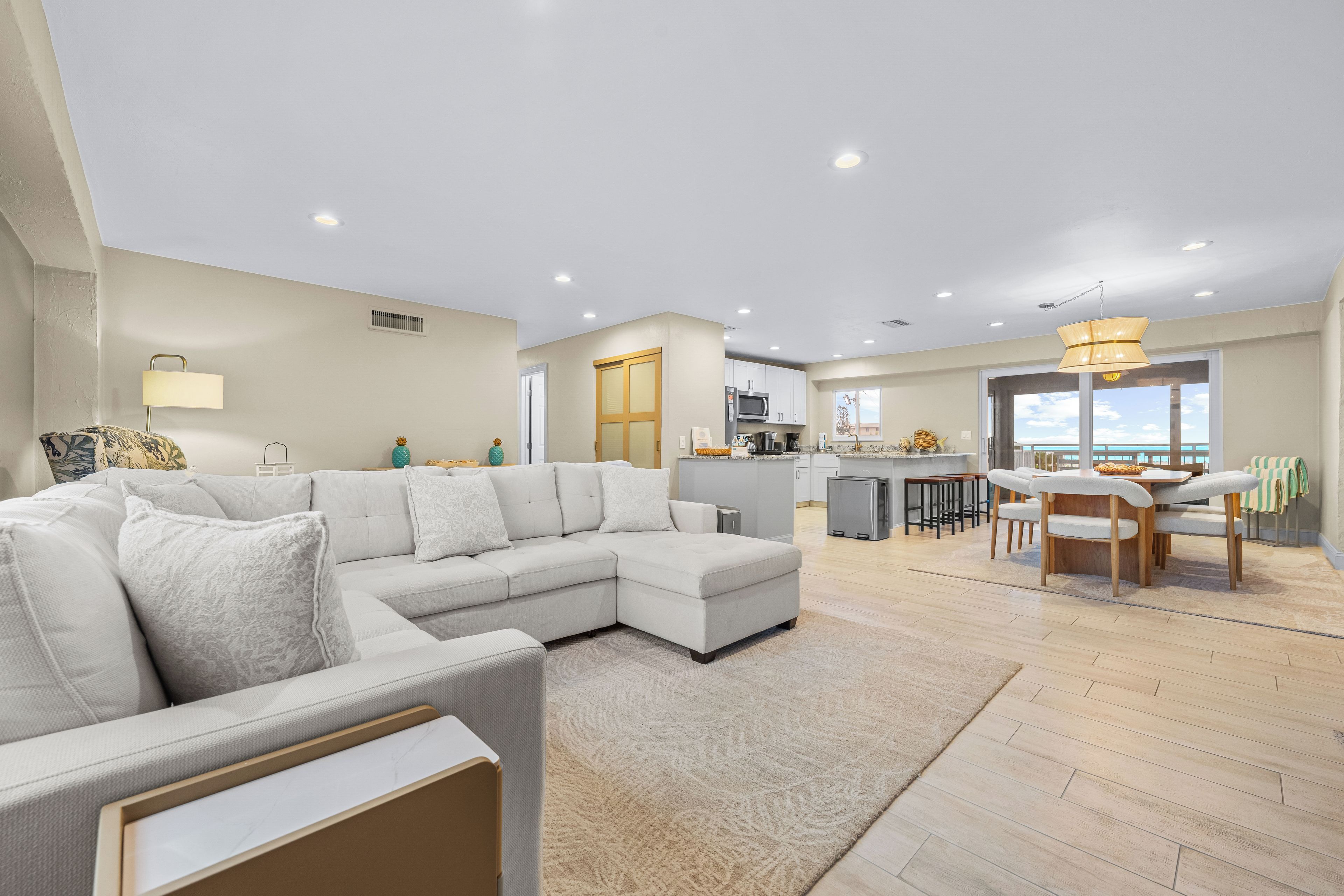 A modern living room with a sectional sofa, a dining area with a table and chairs, and a kitchen with white cabinets and a view to the outside.