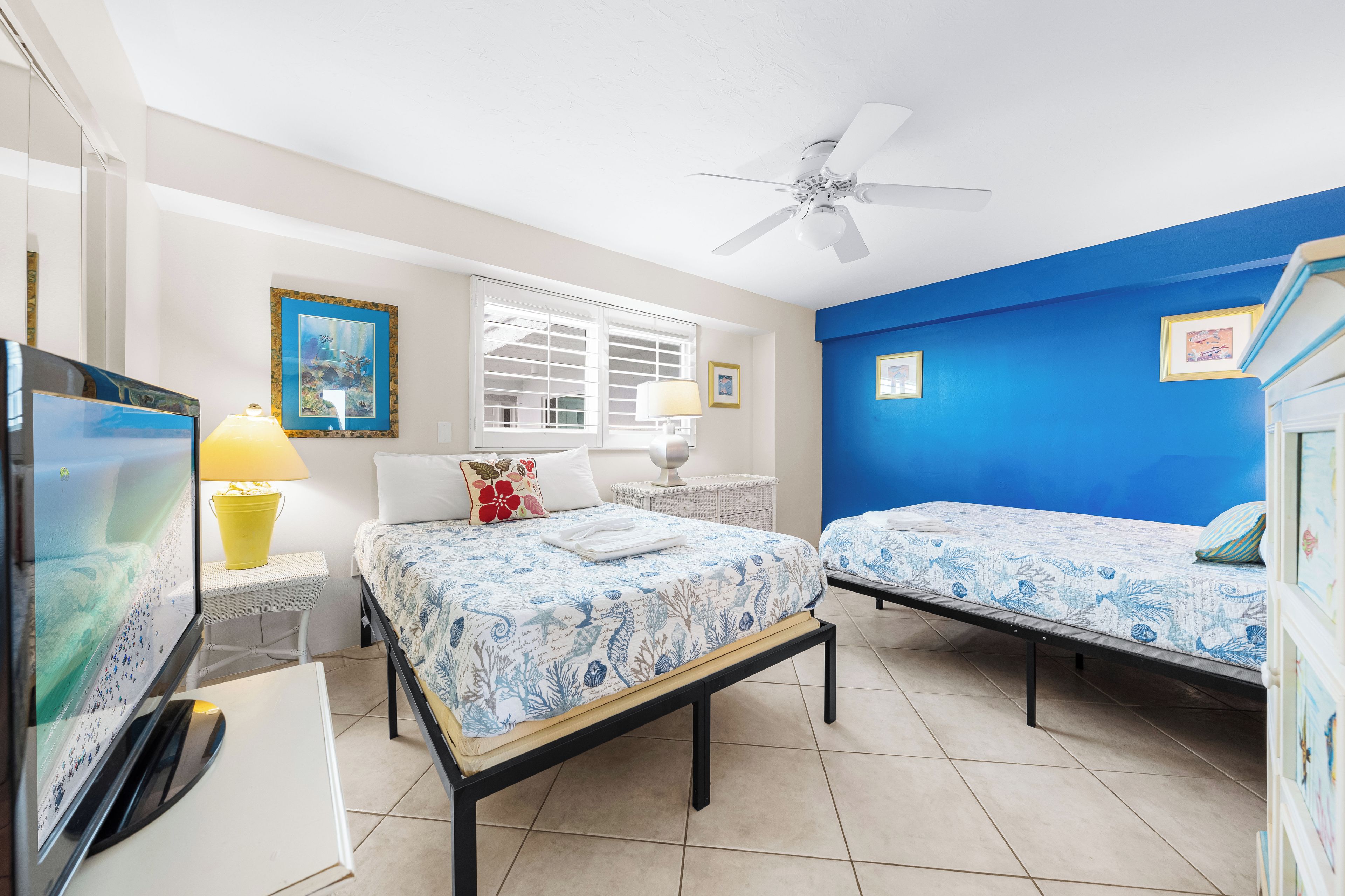 A hotel room with two beds, a ceiling fan, a television, and a window with blinds.