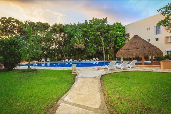 A swimming pool with lounge chairs, a thatched-roof structure, and a building in the background.