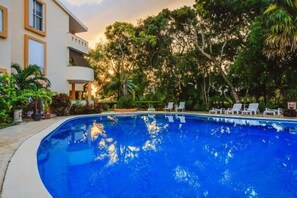 A swimming pool with lounge chairs, surrounded by lush greenery and a building with balconies.