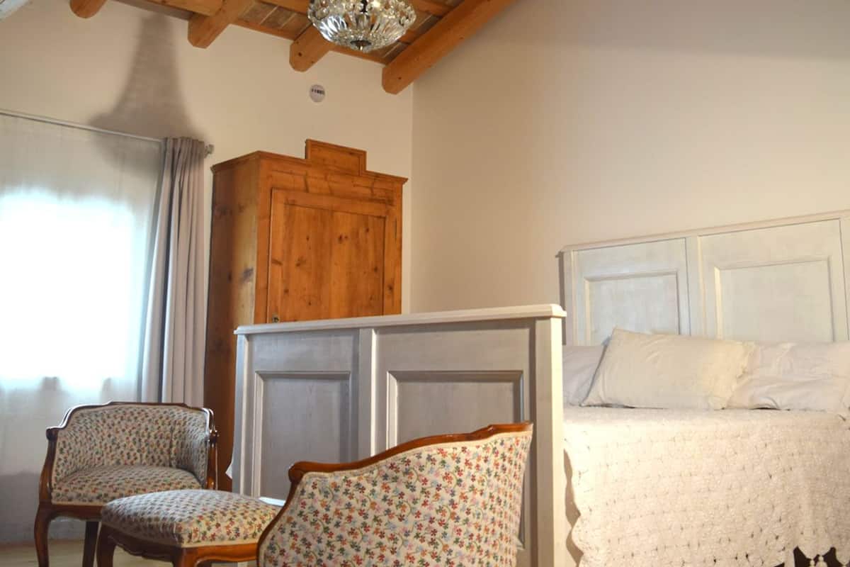 A bedroom with a wooden ceiling, a chandelier, a bed with white bedding, and two floral-patterned chairs.