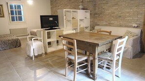 Ein gemütliches Wohnzimmer mit einem Holzesstisch, weißen Stühlen, einem weißen Sofa, einem Fernseher auf einem Schrank und einer Ziegelwand.
