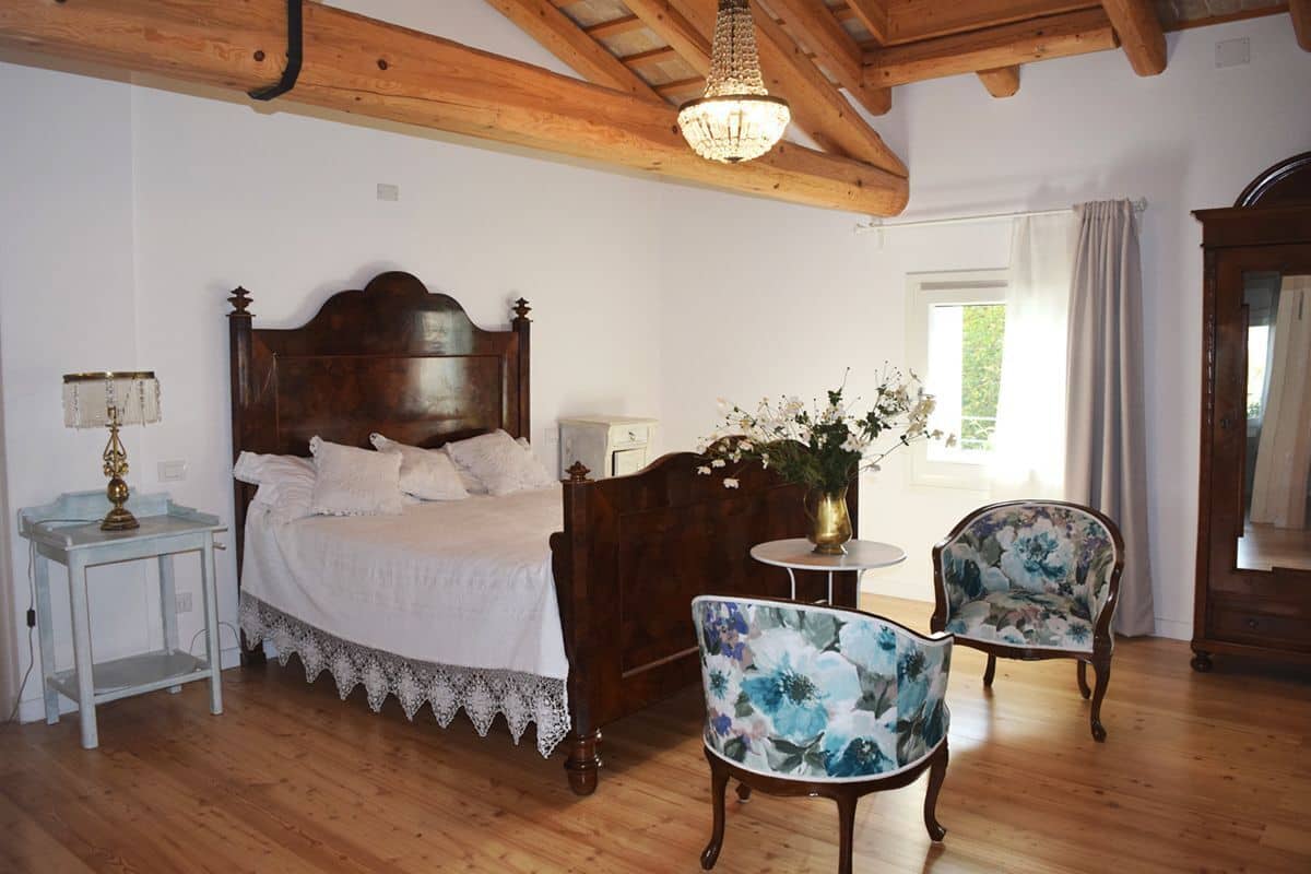 A bedroom with a wooden bed, a nightstand, a lamp, a vase with flowers, two floral chairs, and a wooden dresser.
