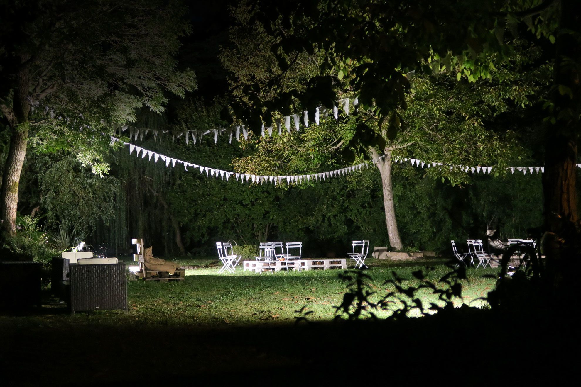 An outdoor seating area with white metal chairs and tables, surrounded by trees and a string of white triangular flags.