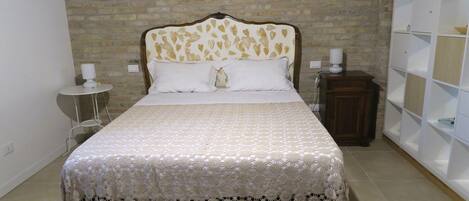 A bed with a patterned bedspread and a decorative headboard, a bedside table with a lamp, and a shelving unit against a brick wall.
