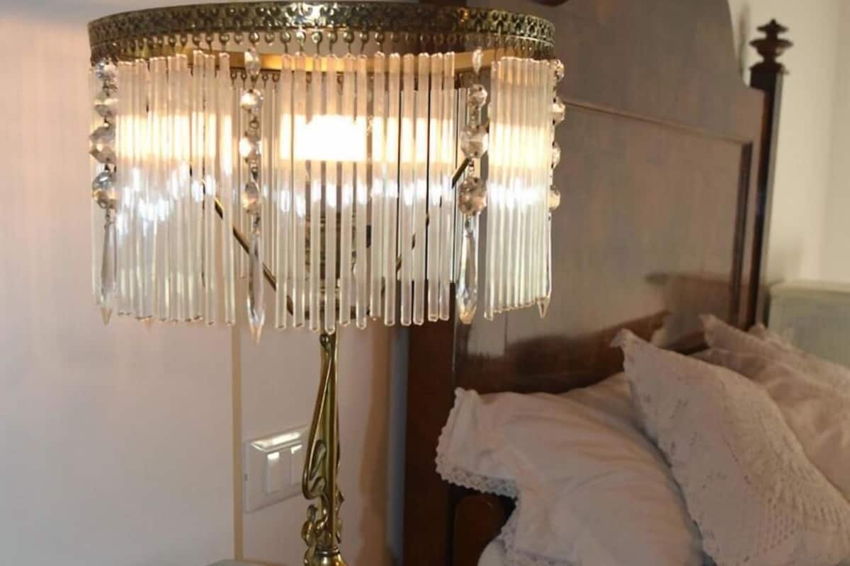 A brass table lamp with a crystal shade on a light blue table in a bedroom with a wooden headboard and white bedding.