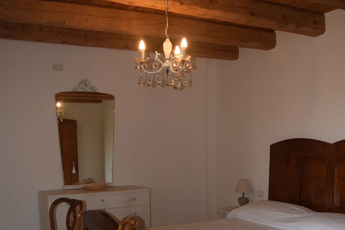 A bedroom with a wooden ceiling, a bed with a white bedspread, a wooden dresser, a chair with a floral cushion, a mirror, and a chandelier.