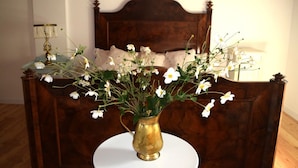 Eine Messingvase mit weißen Blumen auf einem runden Tisch vor einem hölzernen Bett mit Kopfteil.