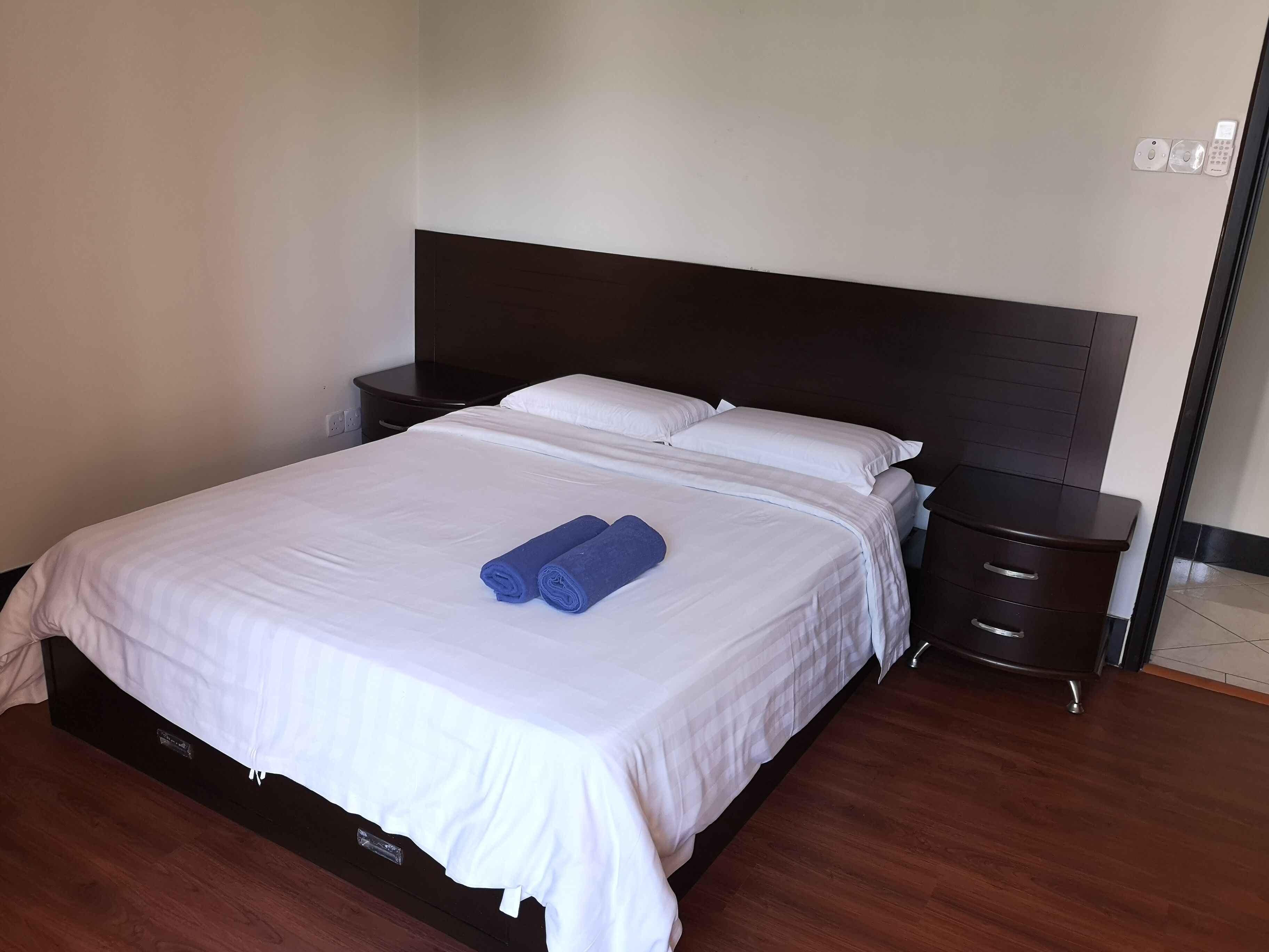 Een net opgemaakt bed met witte lakens en twee opgerolde blauwe handdoeken, een donker houten hoofdbord, een klein nachtkastje met een lamp en een wandcontactdoos.