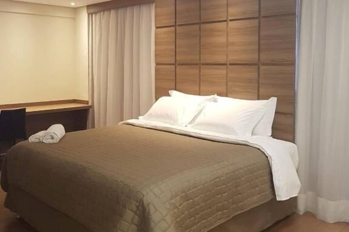 Une chambre d’hôtel avec un grand lit, une tête de lit en bois, un bureau avec un téléphone et une fenêtre avec des rideaux.