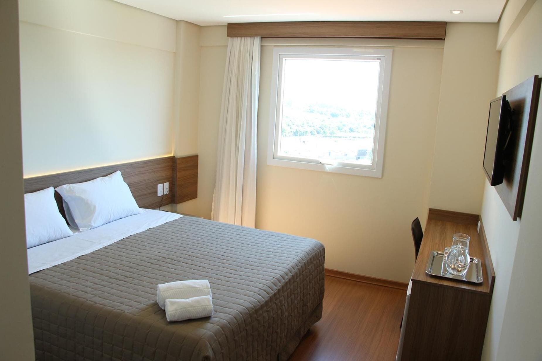 A hotel room with a bed, a bedside table with a glass, and a window with curtains.
