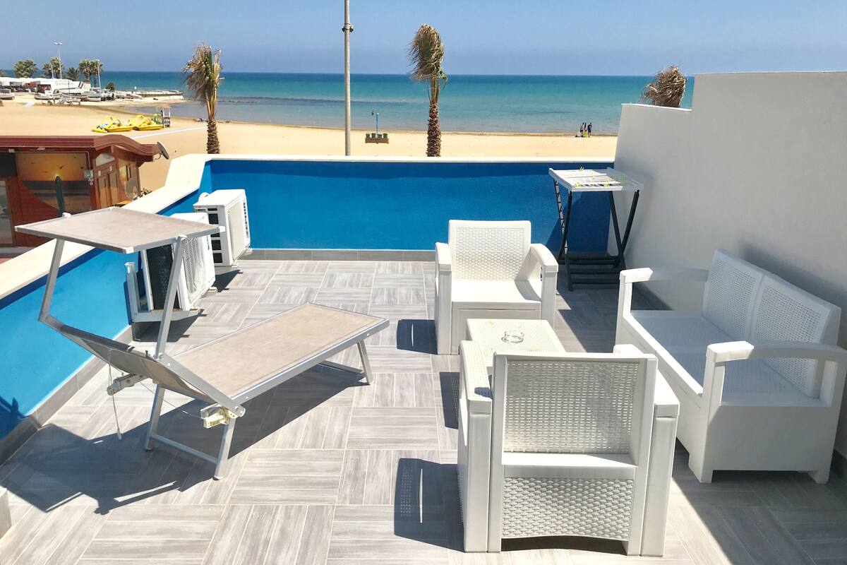 Un espace aménagé au bord de la piscine, avec des chaises longues et une table, offrant une vue sur la plage et la mer.