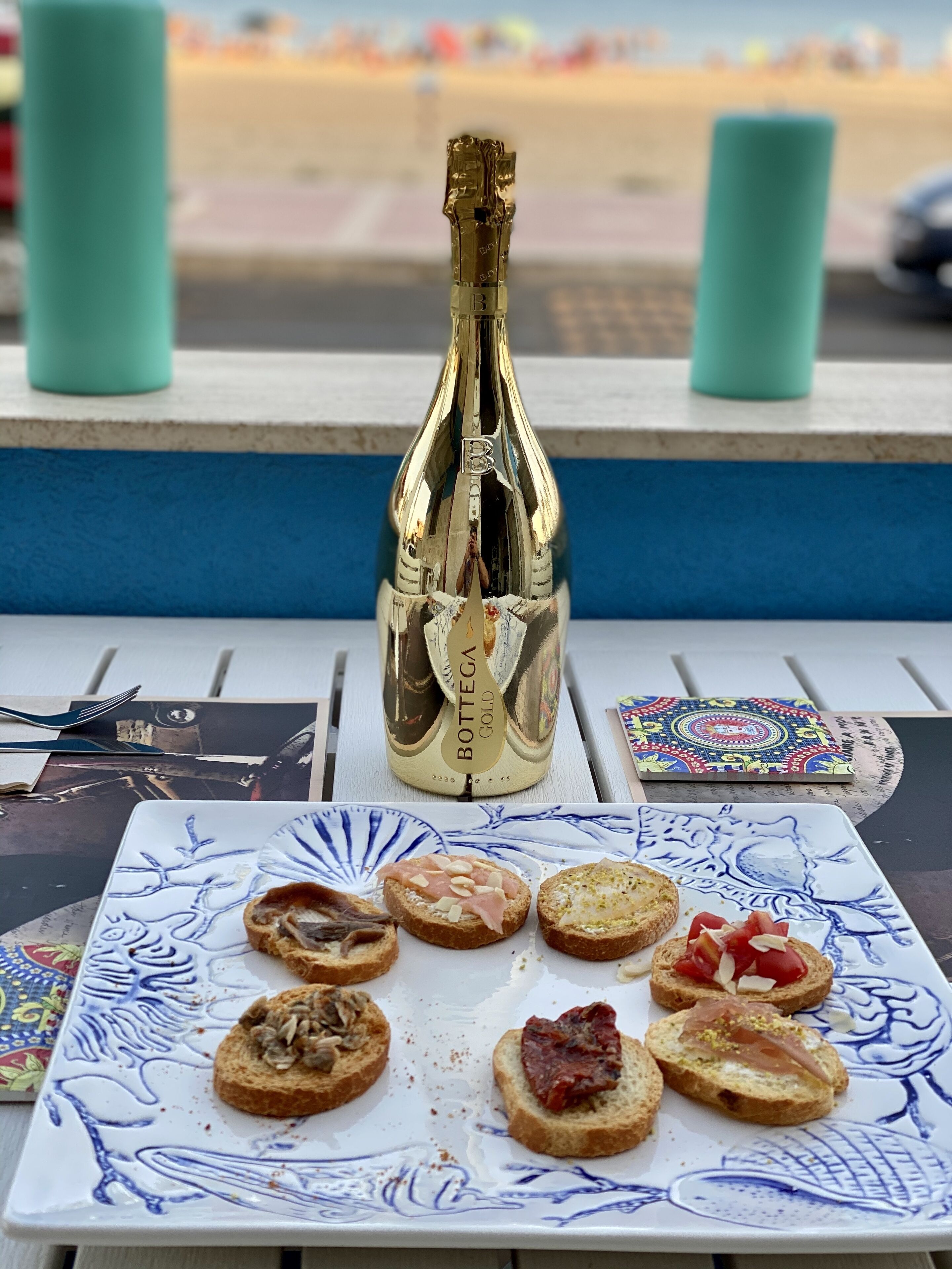 A bottle of Bottega Gold wine, a plate with various appetizers, and a colorful coaster on a table.