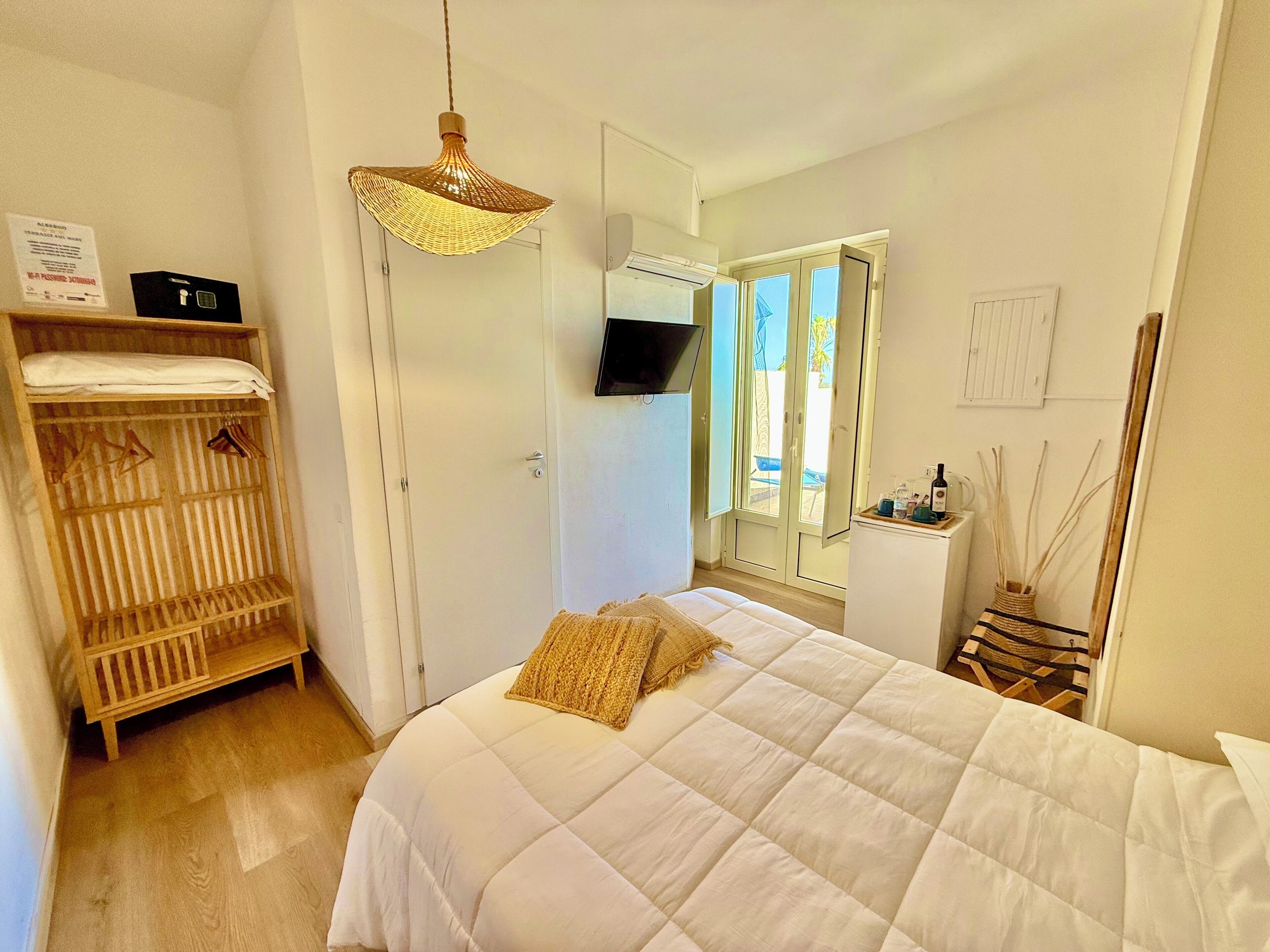 A bedroom with a bed, a wardrobe, a hanging light fixture, a television, and a window with a view of the outdoors.