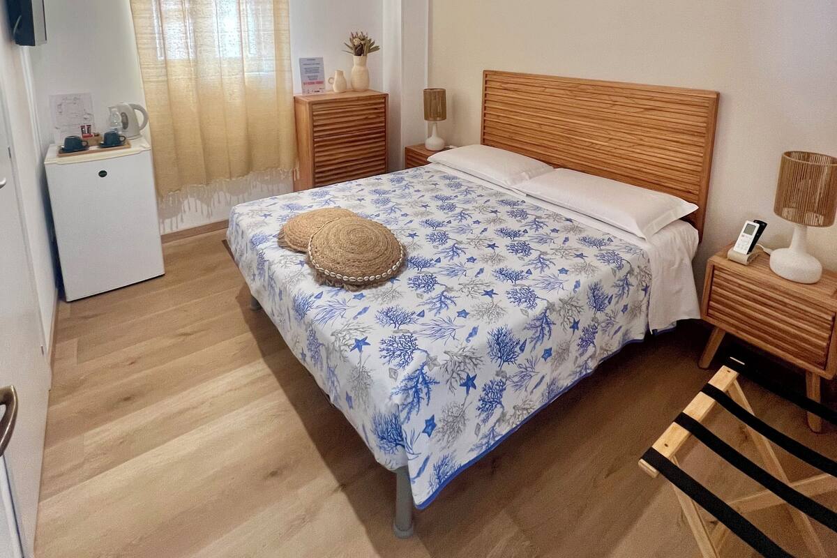 Une chambre avec un lit en bois, une table de chevet avec une lampe, une petite table de chevet avec un vase, un téléviseur fixé au mur et une fenêtre avec des rideaux.