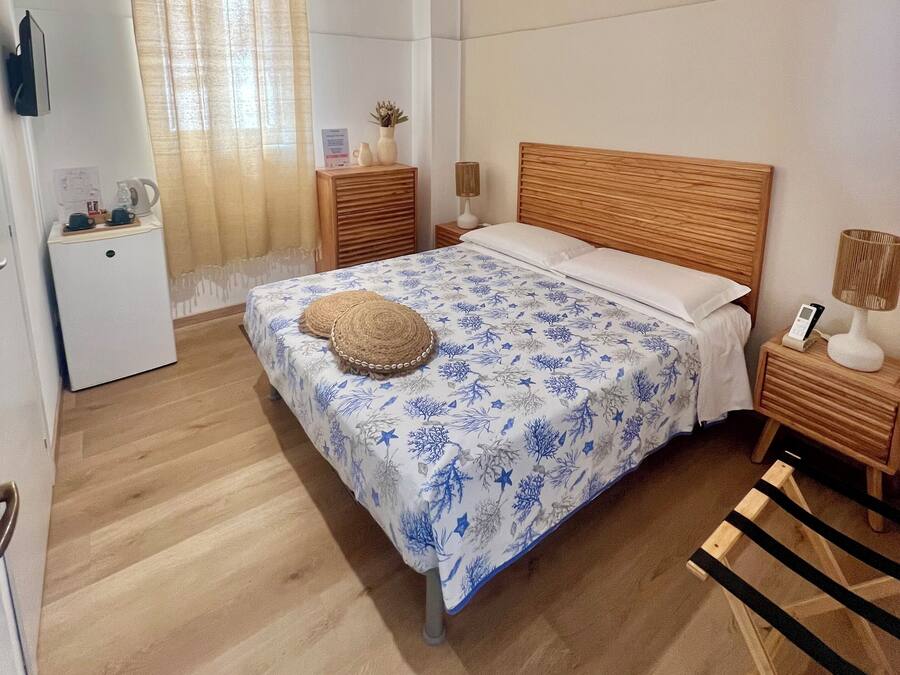 A bedroom with a wooden bed, a bedside table with a lamp, a small bedside table with a vase, a wall-mounted TV, and a window with curtains.