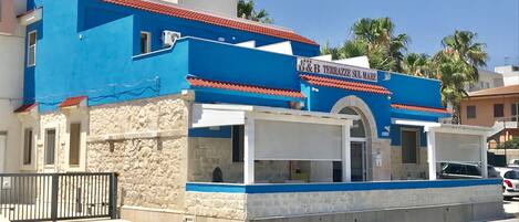 A blue building with a red-tiled roof, a sign reading "B&B TERRAZZE SUL MARE," and a stone wall.