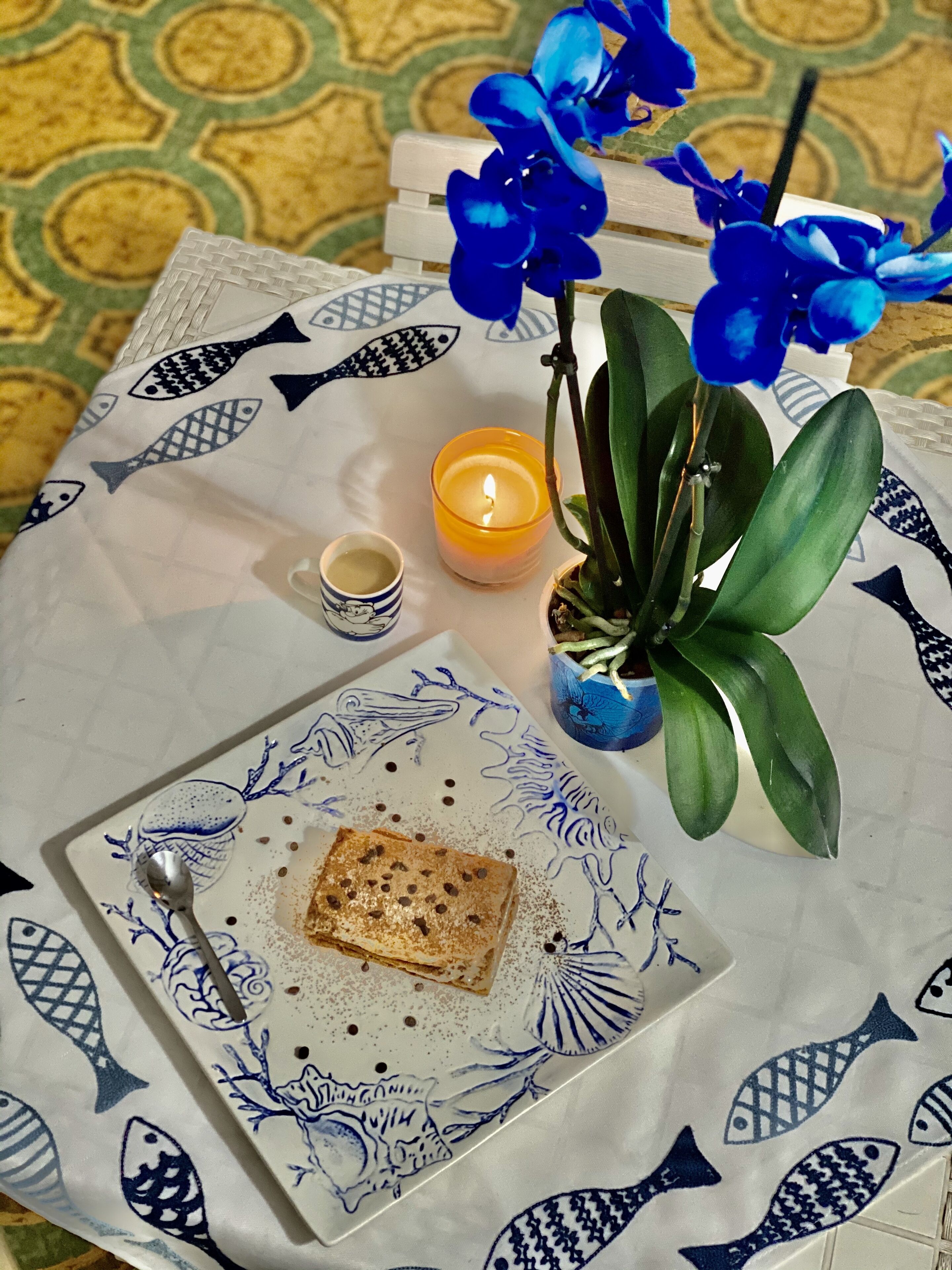 Ein Tisch mit einer Tischdecke mit Fischmuster, eine brennende Kerze, eine kleine Pflanze, eine Tasse und ein Teller mit Kuchen.