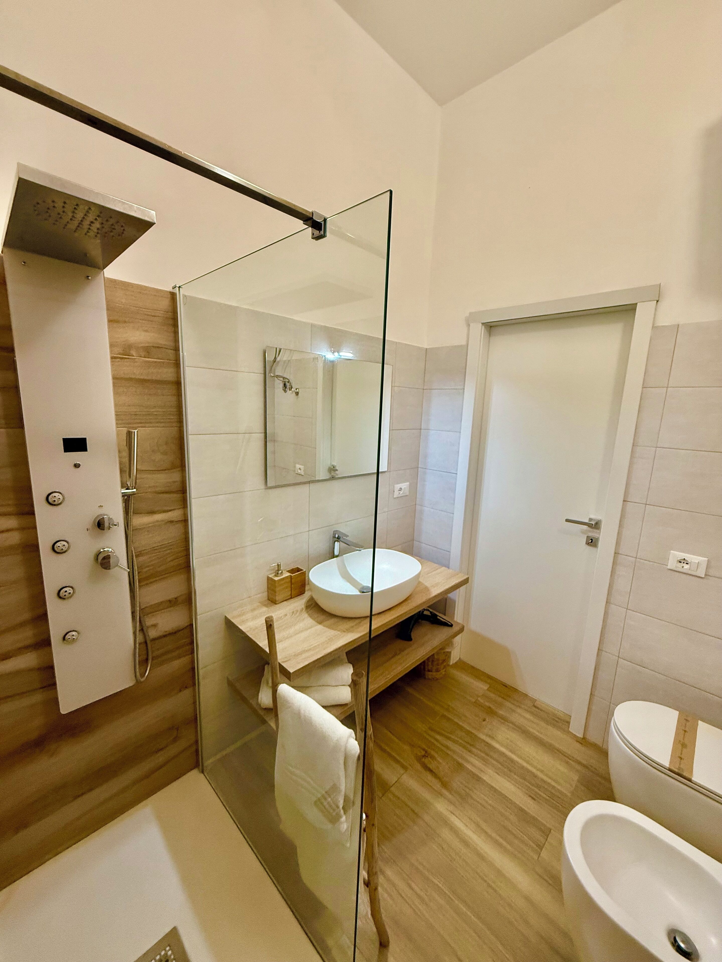 Un baño moderno con ducha envuelta en vidrio, tocador de madera con lavamanos y zona de aseo separada.