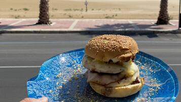 Una mano sosteniendo un plato azul con una hamburguesa y un postre, con vista a una playa con palmeras y un cielo despejado.