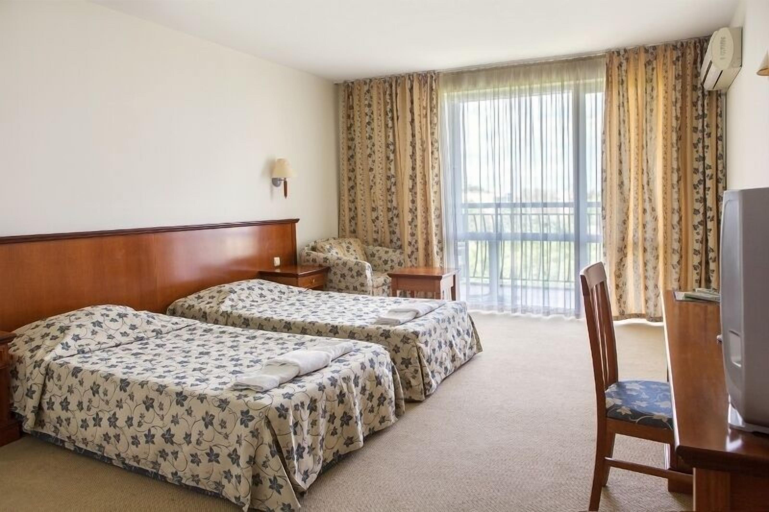 Una habitación de hotel con dos camas, un escritorio, una silla, un televisor y un ventanal grande con cortinas.