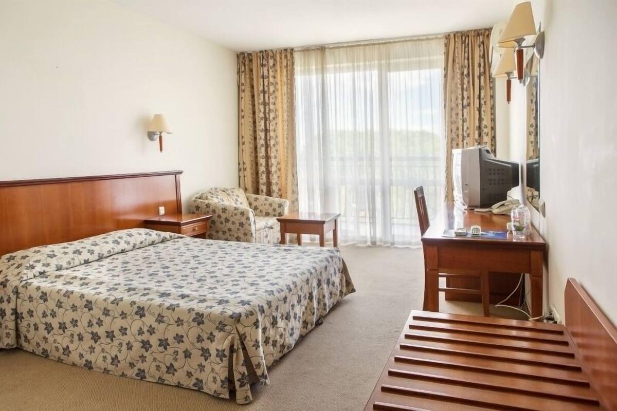 A hotel room with a large bed, a desk with a chair, a small table, a lamp, a television, and a window with curtains.