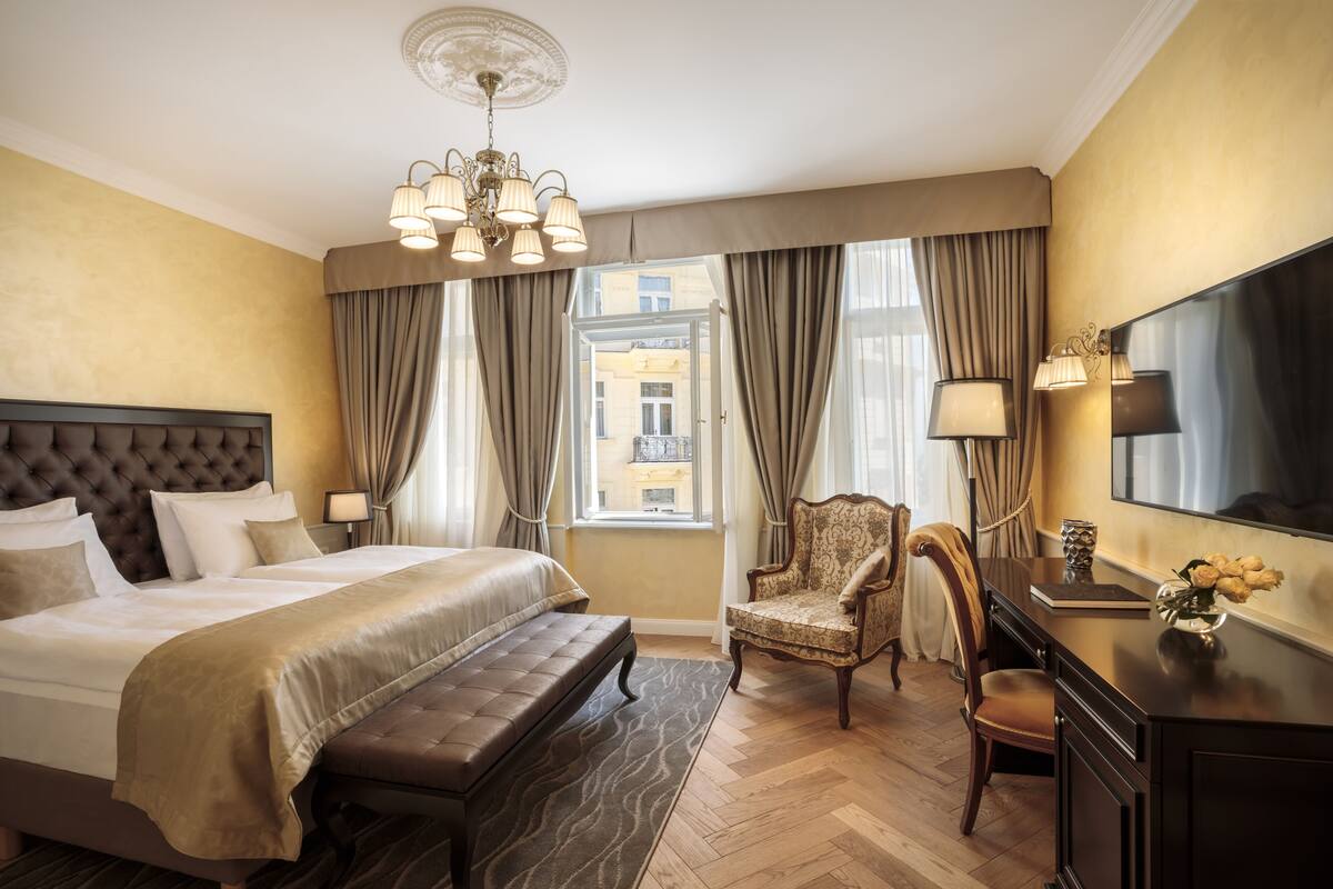 A hotel room with a large bed, a desk with a flat-screen TV, a chair, and a view of the city through the window.