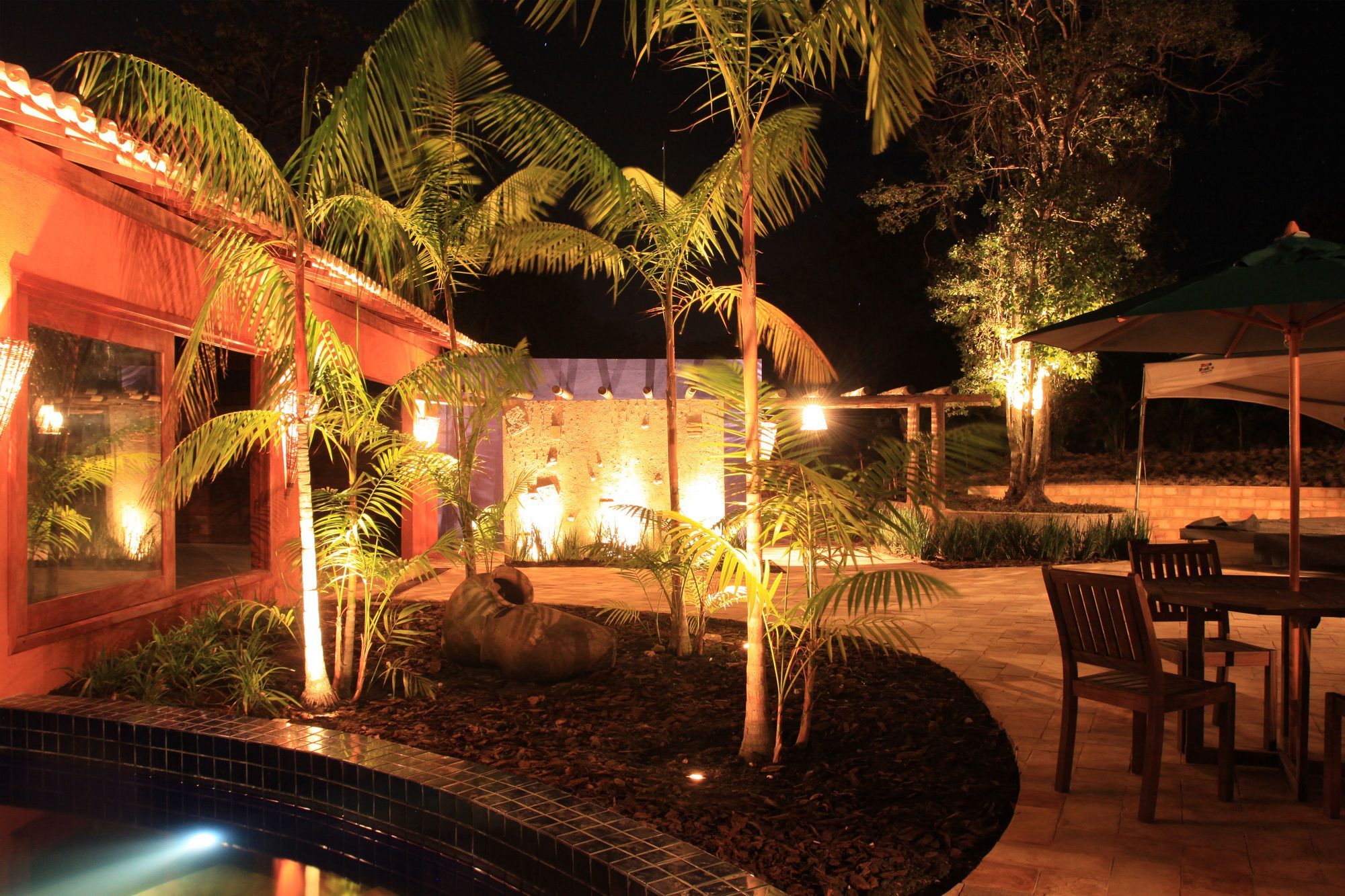 A nighttime outdoor area with a pool, palm trees, and a seating area with a table and chairs.