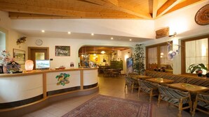 Un espace d’accueil avec un comptoir en bois, une horloge et une grande fenêtre agrémentée de plantes. On y trouve également des coins salon avec des tables et des chaises.