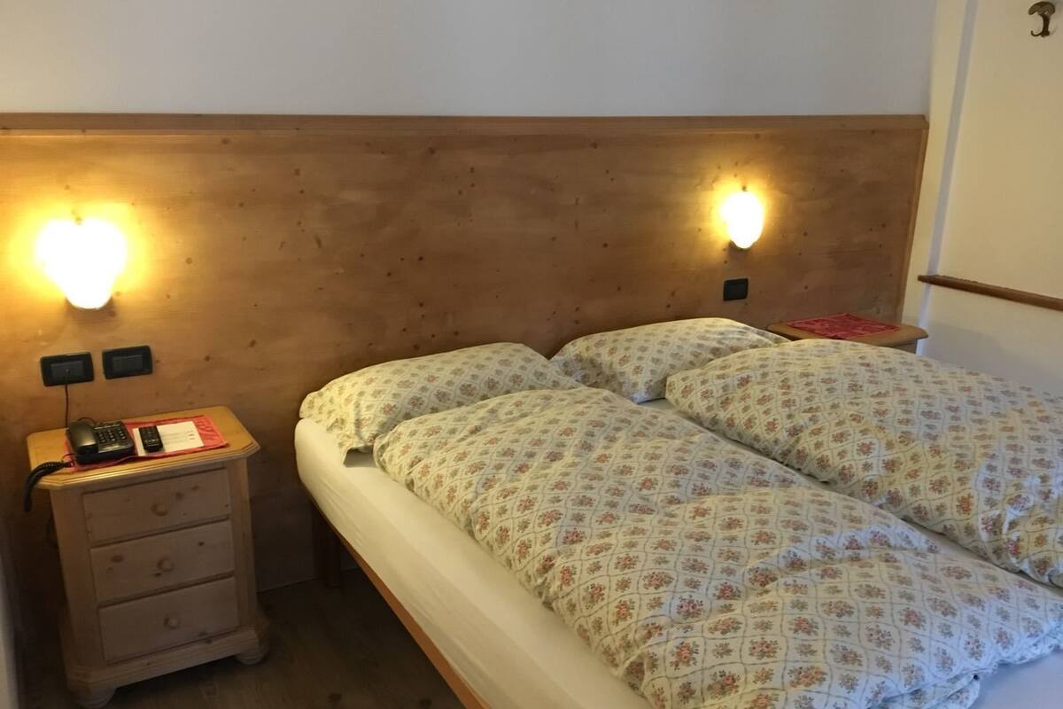 A double bed with floral bedding, a wooden headboard, bedside lamps, a nightstand with a phone and a red object, and a wooden dresser with drawers.