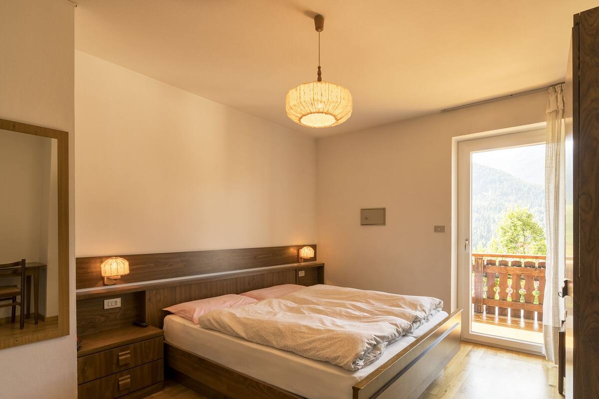 A bedroom with a wooden bed, bedside lamps, a nightstand, and a balcony with a view of the outdoors.