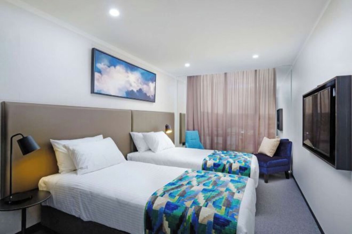 A hotel room with two beds, a TV, a painting, and a window with curtains.