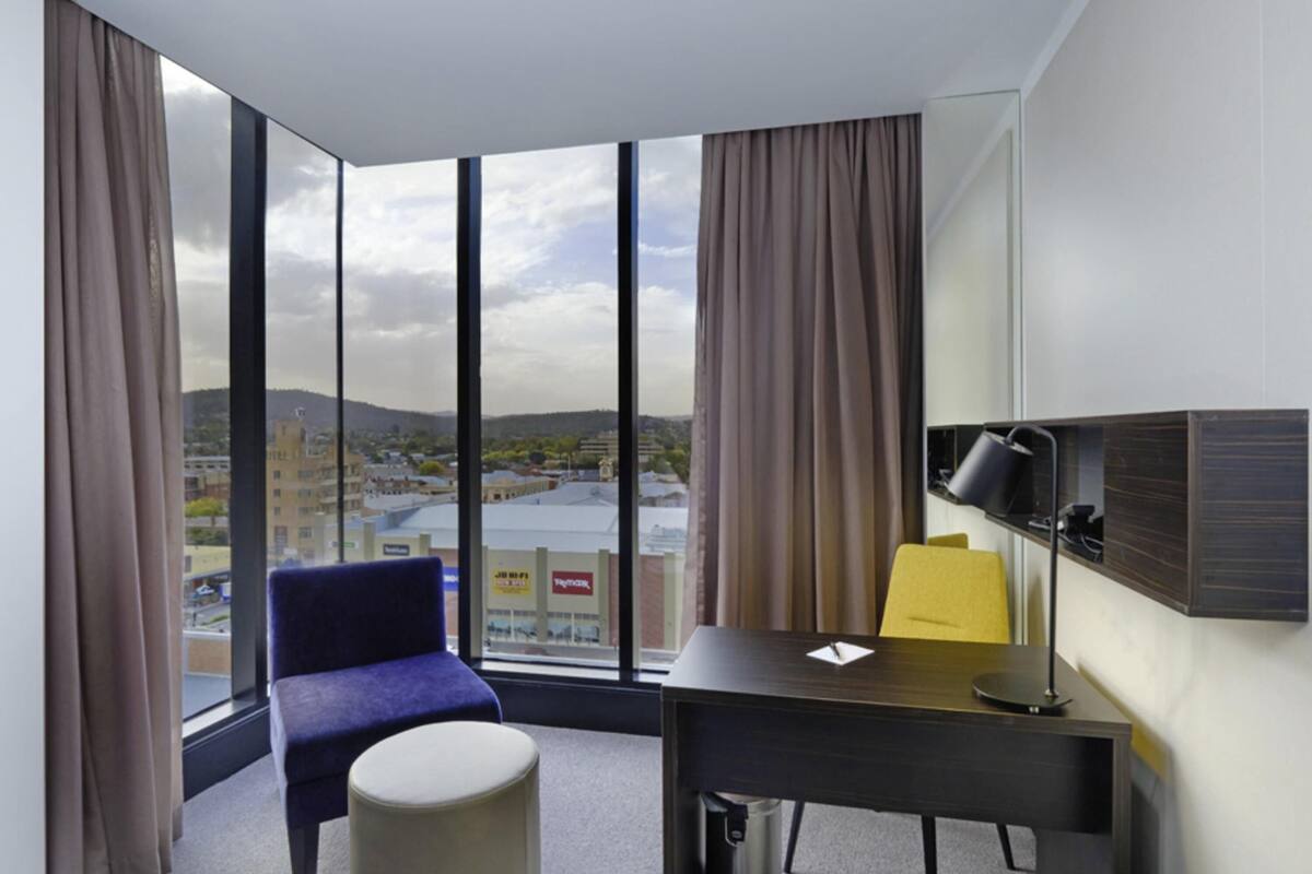Une chambre d’hôtel avec une grande fenêtre, un bureau, une chaise, un petit pouf et un miroir.