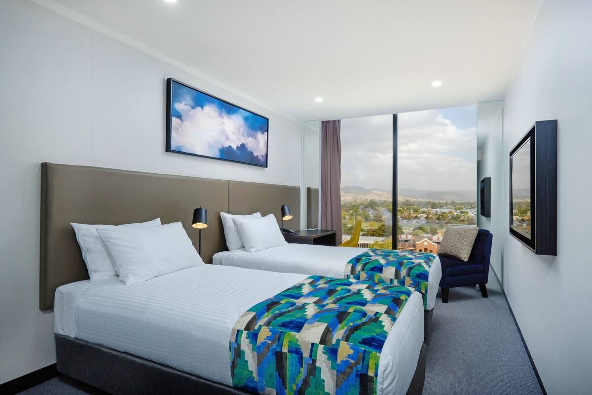 Une chambre d’hôtel avec deux lits, une grande fenêtre donnant sur la ville, un tableau représentant des nuages et une chaise près de la fenêtre.