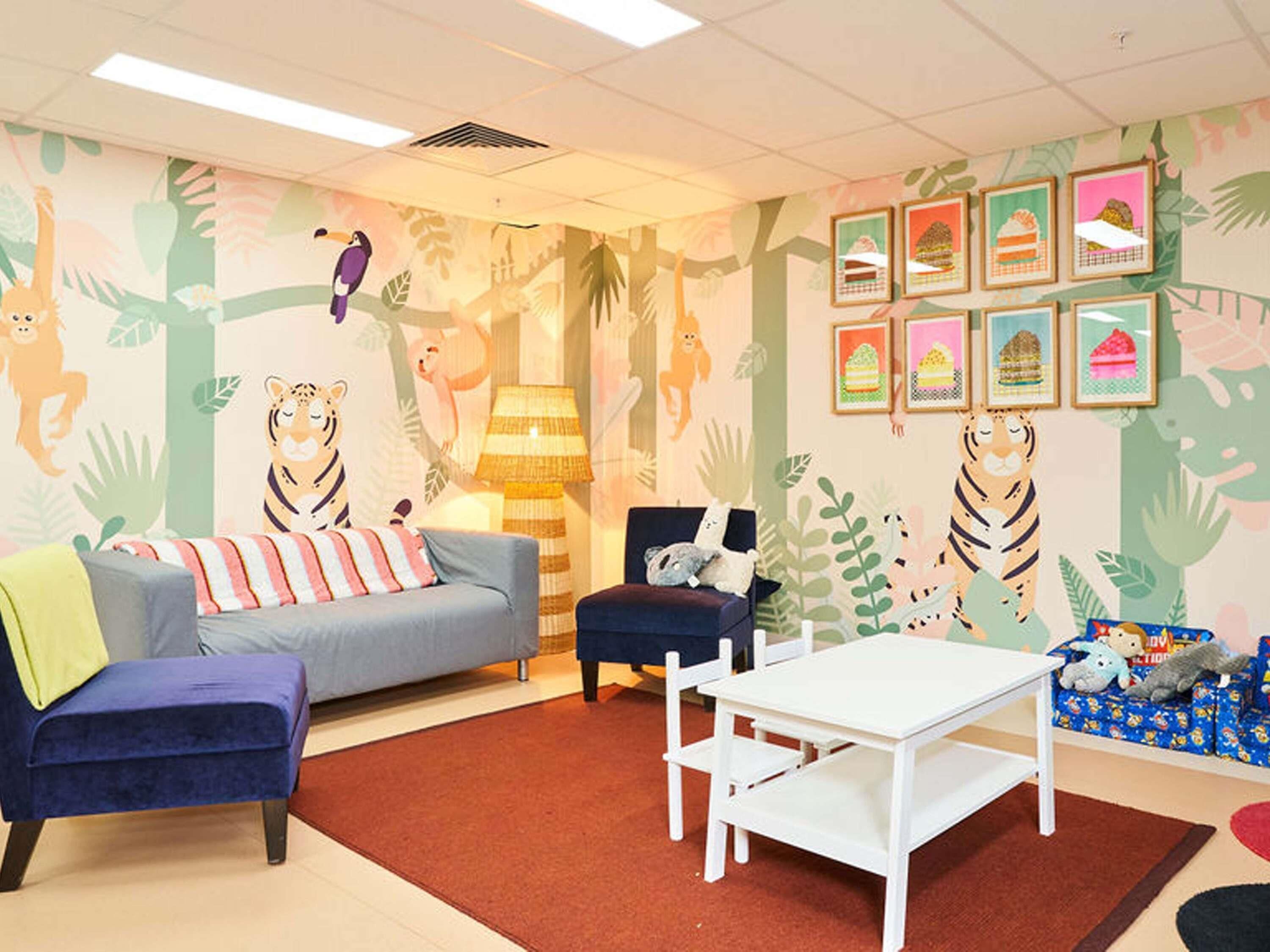 A children's playroom with a sofa, chairs, a small table, and a wall with animal and plant murals.