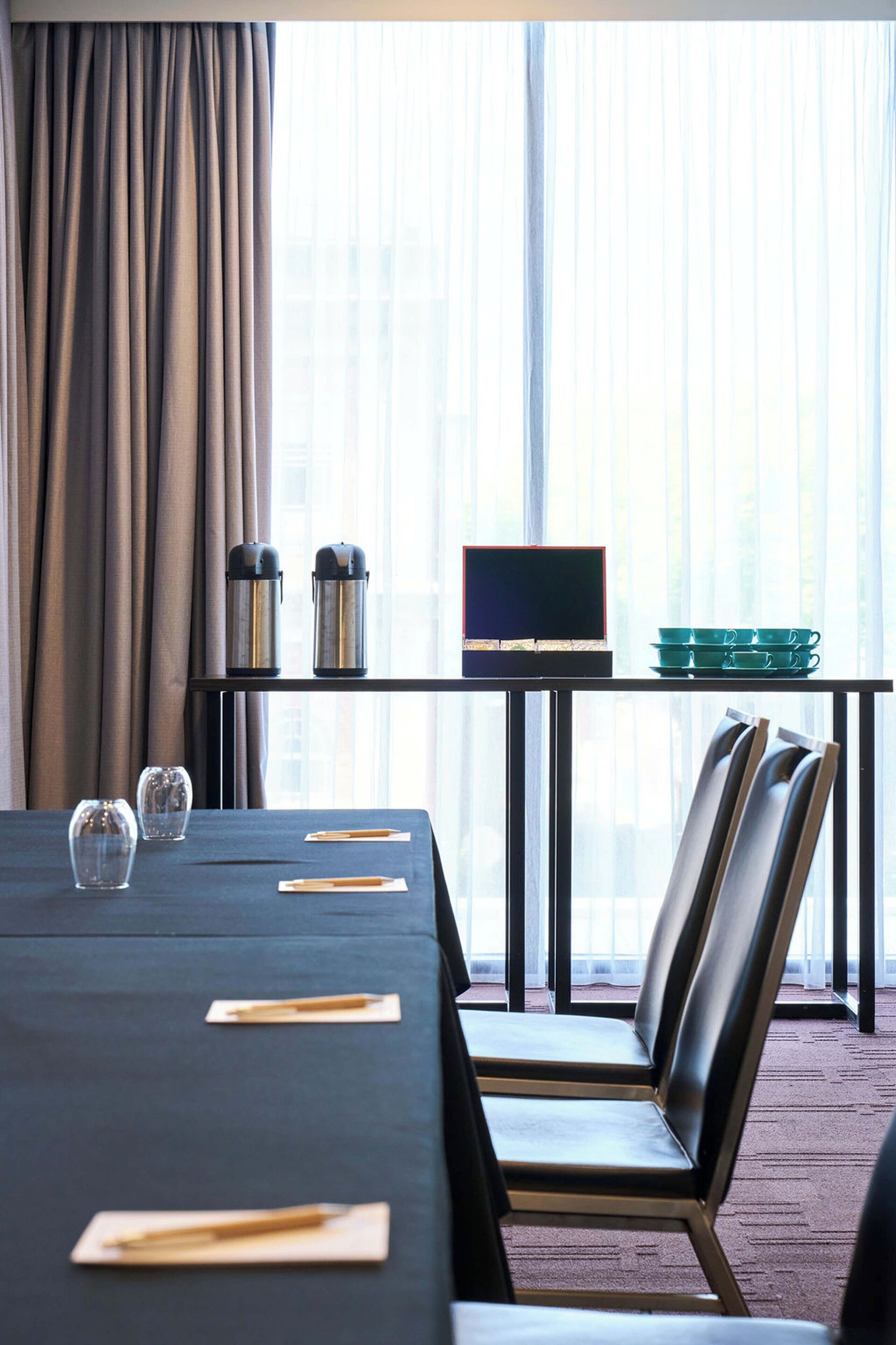 A hotel room with a long table, chairs, a laptop, a coffee maker, and a set of green cups.